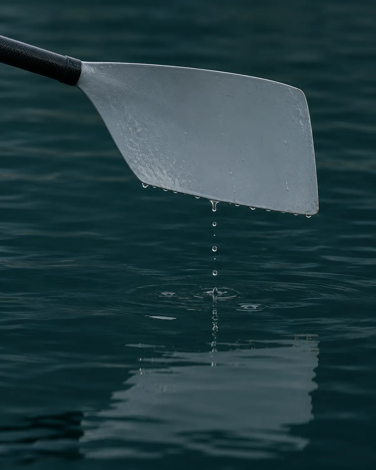 An oar blade held just above still water, drops falling